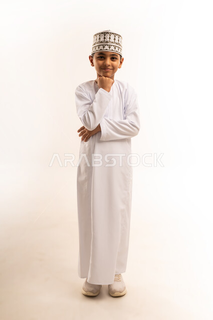 Finding a solution to a problem, contemplation and planning for the future, concentration, absent-mindedness and contemplation, portrait of an Arab Gulf Omani boy wearing a dishdasha and a kummah, standing straight, putting his hand on his chin and looking at the camera with thinking gestures, full-length body image, white background