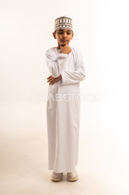 Meditation and planning for the future, finding a solution to a problem, concentration, absent-mindedness and contemplation, portrait of an Arab Gulf Omani boy wearing a dishdasha and a kummah, standing straight, putting his hand on his chin and looking at the camera with thinking gestures, full-length body image, white background