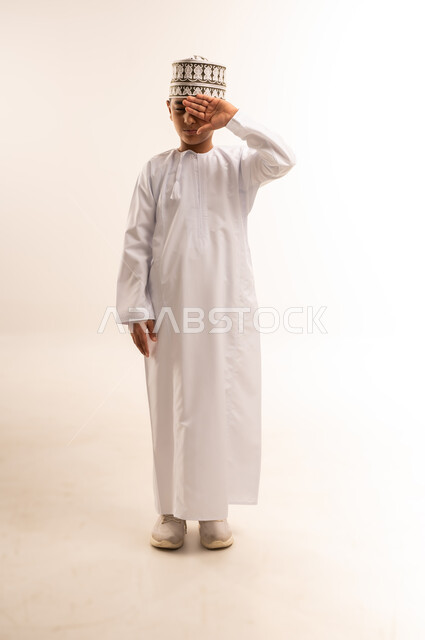 الاحساس بألم في العينين، الشعور بالارهاق وعدم الراحة، مفهوم ضعف البصر، بورتريه لولد عربي خليجي عماني يرتدي الكمة والدشداشة يضع يديه على عينيه بإيماءات النعاس والحاجة الى النوم، تعابير التعب الشديد والصداع، الجسم بالطول الكامل, خلفية بيضاء