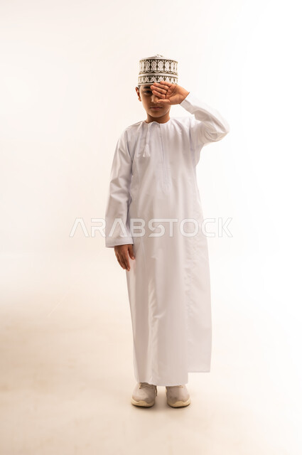 الشعور بالارهاق وعدم الراحة، مفهوم ضعف البصر، الاحساس بألم في العينين، بورتريه لولد عربي خليجي عماني يرتدي الكمة والدشداشة يضع يديه على عينيه بإيماءات النعاس والحاجة الى النوم، تعابير التعب الشديد والصداع، الجسم بالطول الكامل, خلفية بيضاء