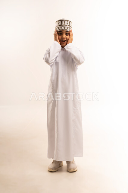 The need for peace and quiet, gestures of disturbance and annoyance from loud noises, portrait of an Arab Gulf Omani boy wearing a dishdasha and a kummah, covering his ears with his fingers to reduce the surrounding noise, full body length, white background