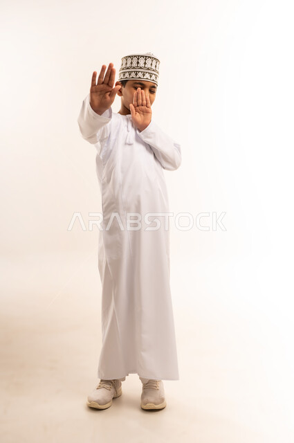 تعابير الذعر والخوف من شيء ما، مد اليدين للأمام محاولة للدفاع عن النفس، بورتريه لولد عربي خليجي عماني يرتدي الدشداشة والكمة تظهر عليه علامات الرعب، ايماءات تدل على طلب النجدة والانقاذ، صورة الجسم بالطول الكامل، خلفية بيضاء