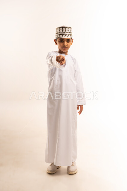 توجيه أصابع الاتهام إلى شخص معين، الإشارة باصبع السبابة إلى الكاميرا بشكل مباشر، التهديد والتوعد بتنفيذ شيء ما، إيماءات التحذير والترهيب، بورتريه لولد عربي خليجي عماني يرتدي الدشداشة والكمة يشير إلى مكان ما محدد، الجسم بالطول الكامل، خلفية بيضاء