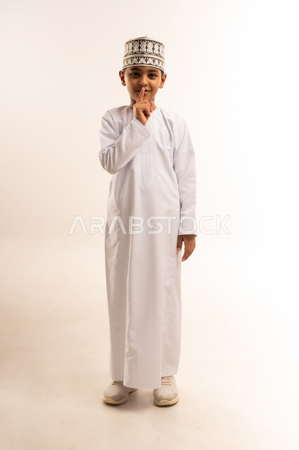 التوقف عن الكلام وخفض الصوت، الهدوء والسكون وعدم الإزعاج، بورتريه لولد عربي خليجي عماني يرتدي الدشداشة والكمة يقف باستقامة ويضع أصبع السبابة على فمه، طلب السكوت والصمت، صورة الجسم بالطول الكامل ، خلفية بيضاء