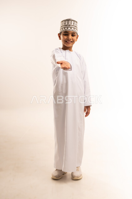 رفع وحمل العناصر، استخدام حاسة اللمس لتحريك العناصر الهولوجراميه، بورتريه لولد عربي خليجي عماني يرتدي الدشداشة والكمة ينظر الى الكاميرا يرفع يده الى الأمام بايماءات حمل شيء، تقديم وعرض عنصر ما، صورة الجسم بالطول الكامل، خلفية بيضاء