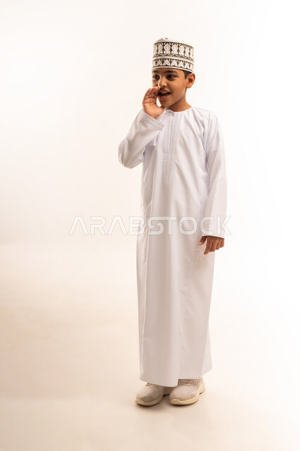 النداء والكلام والتحدث بصوت مرتفع، بورتريه لولد عربي خليجي عماني مبتسم يرتدي الدشداشة و الكمه يقف باستقامة ويقوم بوضع يده على فمه بإيماءات المناداة لشخص ما، دردشة ومحادثات وتواصل اجتماعي، الطول الكامل للجسم، خلفية بيضاء