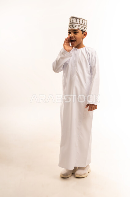 الكلام والنداء والتحدث بصوت مرتفع، بورتريه لولد عربي خليجي عماني مبتسم يرتدي الدشداشة و الكمه يقف باستقامة ويقوم بوضع يده على فمه بإيماءات المناداة لشخص ما، دردشة ومحادثات وتواصل اجتماعي، الطول الكامل للجسم، خلفية بيضاء