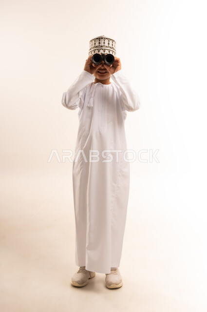 رؤية الأشياء البعيدة باستخدام التلسكوب، حب الاستكشاف والمغامرة، التخطيط للأهداف والطموحات المستقبلية، بورتريه لولد عربي خليجي عماني يرتدي الدشداشة والكمة يقف باستقامة ويقوم بالنظر من خلال المنظار، الطول الكامل للجسم، خلفية بيضاء