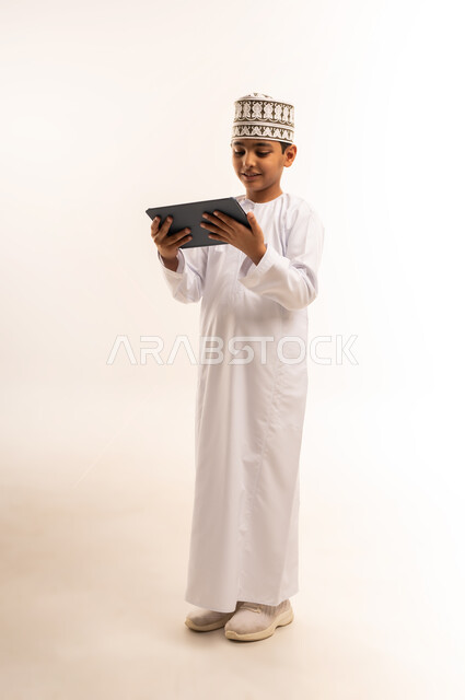 مشاهدة البرامج الترفيهيه المفضلة بإيماءات السعادة والسرور، بورتريه لولد عربي خليجي عماني مبتسم يرتدي الكمة والدشداشة يحمل بيده جهاز التابليت، استخدام التقنية الحديثة في تزويد الأجيال بالمعلومات المفيدة، صورة الجسم بالطول الكامل، خلفية بيضاء