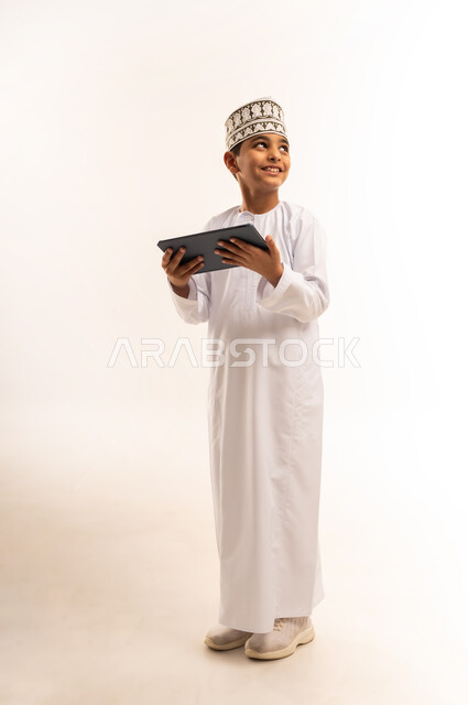 استخدام التقنية الحديثة في تزويد الأجيال بالمعلومات المفيدة، مشاهدة البرامج الترفيهيه المفضلة بإيماءات السعادة والسرور، بورتريه لولد عربي خليجي عماني مبتسم يرتدي الكمة والدشداشة يحمل بيده جهاز التابليت، صورة الجسم بالطول الكامل، خلفية بيضاء