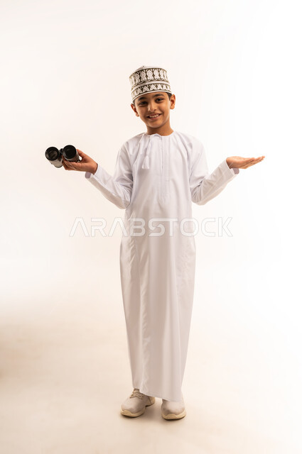 رؤية الأشياء البعيدة، التخطيط المستقبلي للأهداف والطموحات، حب الاستكشاف والمغامرة، بورتريه لولد عربي خليجي عماني يرتدي الدشداشة والكمة يمسك منظار ثنائي العينين ينظر إلى الكاميرا بإيماءات السرور، التفكير والتطلع إلى المستقبل، الجسم بالطول الكامل، خلفية بيضاء