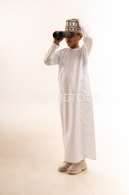 التخطيط المستقبلي للأهداف والطموحات، حب الاستكشاف والمغامرة، بورتريه لولد عربي خليجي عماني يرتدي الدشداشة والكمة ينظر من خلال منظار ثنائي العينين إلى شيء ما بإيماءات الدهشة، رؤية الأشياء البعيدة، التفكير والتطلع إلى المستقبل، الجسم بالطول الكامل، خلفية بيضاء
