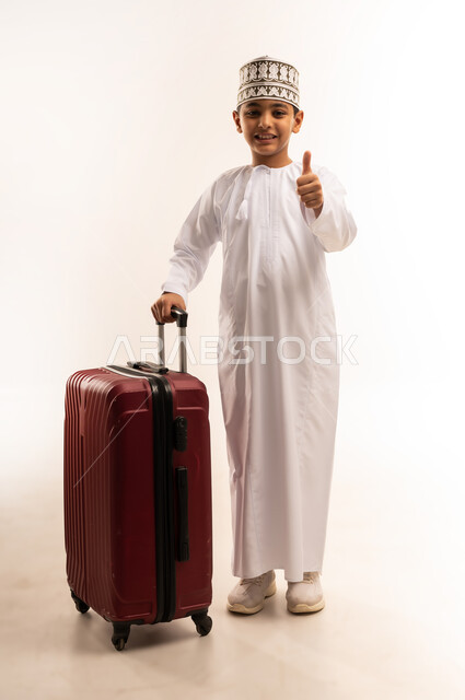 علامة الجودة والامتياز، القيام بجولة ترفيهية، بورتريه لولد عربي خليجي عماني يرتدي الدشداشة والكمة يقف بجانب حقيبة الأمتعة يرفع اصبع الابهام بايماءات القبول والاعجاب، الفرح لقضاء رحلة صيفية ممتعة، مفهوم السياحة في إجازة الصيف، الجسم بالطول الكامل، خلفية بيضاء