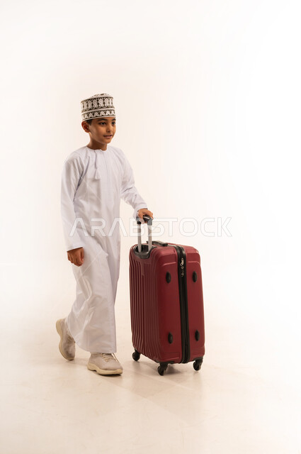 المشي والذهاب إلى مكان ما، القيام بجولة ترفيهية، بورتريه لولد عربي خليجي عماني يرتدي الدشداشة والكمة يقوم بجر حقيبة الأمتعة، الفرح لقضاء رحلة صيفية ممتعة، مفهوم السياحة في إجازة الصيف، الجسم بالطول الكامل، خلفية بيضا