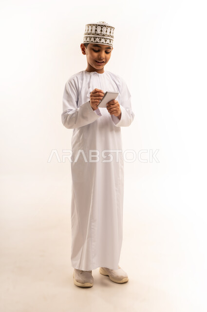 Reviewing lessons and solving duties, the concept of notes notes, diligence and diligence to reach success, portrait for an Arab Gulf Arab boy who wears a dishdasha and the punch carrying a small pen and book that writes, the body is full, white background
