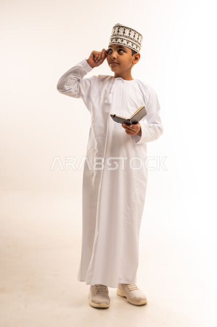 مراجعة الدروس وحل الواجبات، الجد والاجتهاد للوصول إلى النجاح، بورتريه لولد عربي خليجي عماني يرتدي الدشداشة والكمة يحمل قلم ودفتر صغير يقوم بالكتابة ينظر إلى الاعلى بإيماءات التفكير ومحاولة التذكر، مفهوم تدوين الملاحظات، الجسم بالطول الكامل, خلفية بيضاء