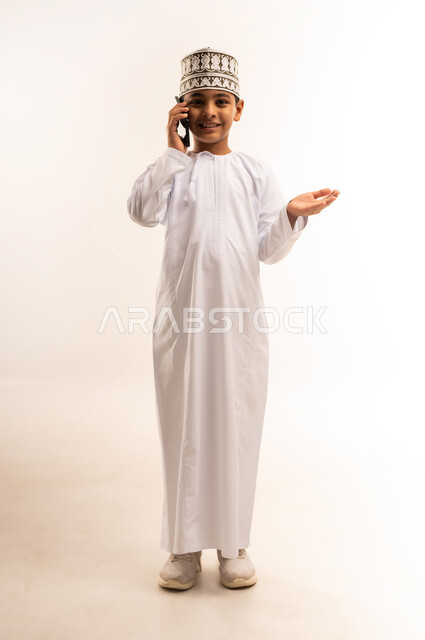 استخدام جهاز تقني حديث ومتطور، الدردشة والتواصل مع الاهل والاصدقاء عبر الجوال، بورتريه لولد عربي خليجي عماني يرتدي الدشداشه والكمه يمسك الهاتف المحمول يقوم باجراء مكالمة هاتفية, الجسم بالطول الكامل، خلفيه بيضاء