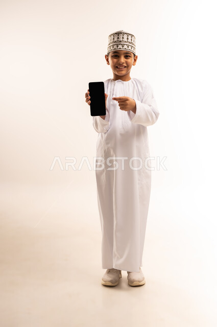 عرض شاشة سوداء فارغة، استخدام جهاز تقني حديث ومتطور، الدردشة والتواصل مع الاهل والاصدقاء عبر الجوال، بورتريه لولد عربي خليجي عماني يرتدي الدشداشه والكمه يمسك الهاتف المحمول ينظر إلى الكاميرا بإيماءات السرور، الجسم بالطول الكامل، خلفيه بيضاء