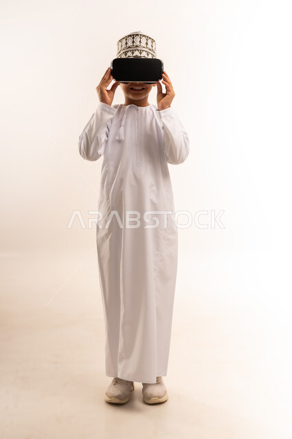 Simulating the imaginary world, fun and entertainment by watching through virtual reality glasses, portrait of an Arab Gulf Omani boy wearing a dishdasha and a kimono, looking through 3D glasses (VR) with gestures of pleasure, using a modern and advanced technical device, full-length body, white background
