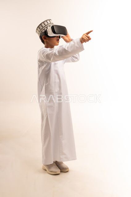 Pointing with index finger at something, simulating the imaginary world, enjoying watching through virtual reality glasses, portrait of an Arab Gulf Omani boy wearing a dishdasha and a kilt looking through 3D glasses (VR), using a modern and advanced technical device, full-length body, white background