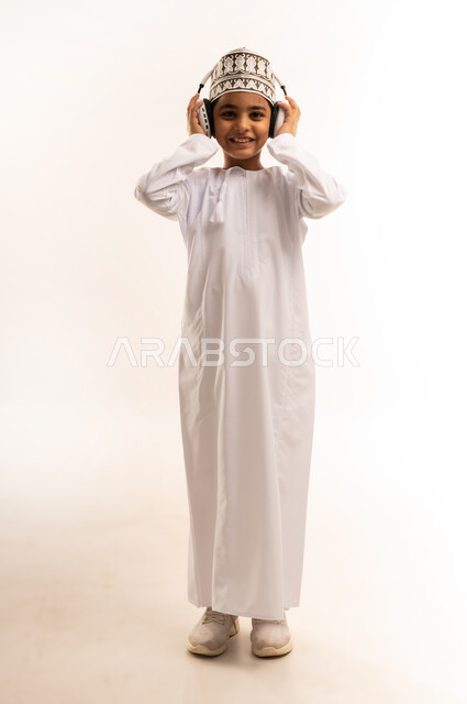 التفاعل مع الأنغام الموسيقية، الانصات والاستماع للأغاني باستخدام الهيدفون، بورتريه لولد عربي خليجي عماني يرتدي الدشداشة والكمة يستمع إلى الموسيقى من خلال سماعة الرأس ينظر إلى الكاميرا بإيماءات السرور والاندماج، الجسم بالطول الكامل، خلفية بيضاء