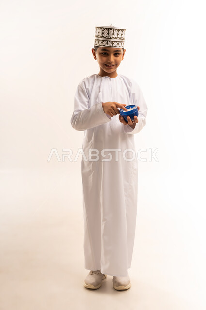 الاهتمام بالمظهر الخارجي، بورتريه لفتى عربي خليجي عماني يرتدي الدشداشة والكمة يمسك بيده كريم الترطيب، مفهوم العناية بنضارة وتحسين البشرة، استخدام كريمات ومستحضر تجميلي، الجسم بالطول الكامل، خلفية بيضاء