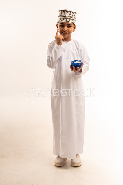 الاهتمام بالمظهر الخارجي، بورتريه لفتى عربي خليجي عماني يرتدي الدشداشة والكمة يمسك بيده كريم الترطيب يقوم بوضعه على خده، مفهوم العناية بنضارة وتحسين البشرة، استخدام كريمات ومستحضر تجميلي، الجسم بالطول الكامل، خلفية بيضاء
