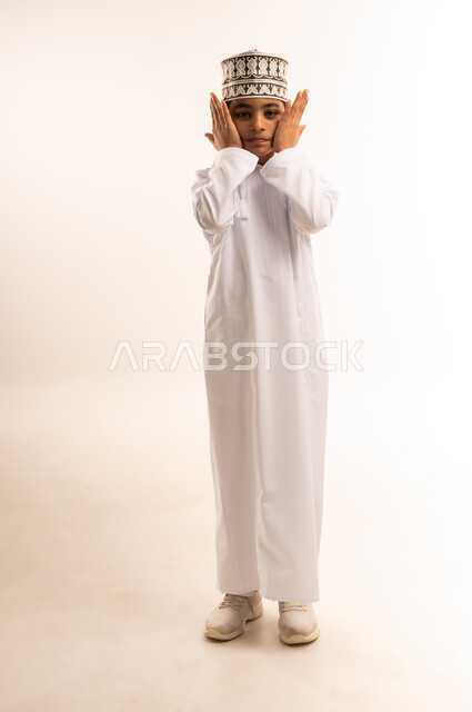 الاهتمام بالمظهر الخارجي، بورتريه لفتى عربي خليجي عماني يرتدي الدشداشة والكمة يضع يديه على وجهه يقوم بدهن شيء ما على خديه، مفهوم العناية بالوجه، الجسم بالطول الكامل، خلفية بيضاء
