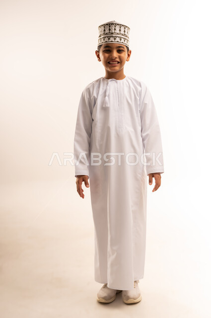 مفهوم الاناقة والوسامة، الاهتمام بالمظهر الخارجي, بورتريه لولد عربي خليجي عماني يرتدي الدشداشة و الكمة يقف باستقامة ينظر إلى الكاميرا بإيماءات السرور والسعادة، صورة الجسم بالطول الكامل, خلفية بيضاء