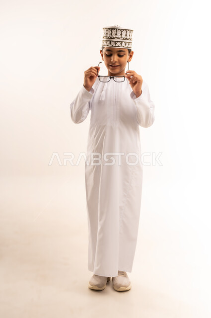 مفهوم الاناقة والوسامة، الاهتمام بالمظهر الخارجي, بورتريه لولد عربي خليجي عماني يرتدي الدشداشة و الكمة يقوم بارتداء النظارات الطبية، النظر إلى الكاميرا بإيماءات السرور والسعادة، صورة الجسم بالطول الكامل, خلفية بيضاء