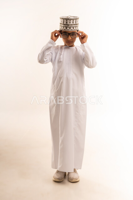 الاهتمام بالمظهر الخارجي, بورتريه لولد عربي خليجي عماني يرتدي الدشداشة و الكمة يقوم بارتداء النظارات الطبية، مفهوم الاناقة والوسامة، النظر إلى الكاميرا بإيماءات السرور والسعادة، صورة الجسم بالطول الكامل, خلفية بيضاء