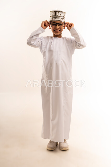 Looking at the camera with gestures of pleasure and happiness, interest in appearance, portrait of an Arab Gulf Omani boy wearing a dishdasha and a kummah wearing medical glasses, concept of elegance and handsomeness, full-length body image, white background