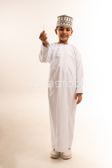 التأكد من نظافة الأسنان بعد تناول الطعام، المحافظة على ابتسامة ناصعة البياض، بورتريه لفتى عربي خليجي عماني يرتدي الدشداشة والكمة يمسك بيده الخيط السني, مفهوم العناية بصحة الفم، الاهتمام بالأناقة والمظهر الخارجي، الجسم بالطول الكامل، خلفية بيضاء