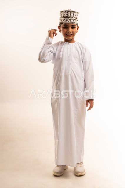 روتين نظافة الأذنين اليومية، العناية والاهتمام بصحة الأذن، استخدام أعواد ونكاشات القطن، بورتريه لفتى عربي خليجي عماني يرتدي الدشداشة والكمة يقوم وضع نكاشة قطنية داخل أذنه، الاهتمام المظهر الخارجي، الجسم بالطول الكامل، خلفية بيضاء