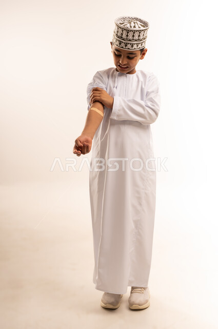 التضميد الصحيح للجراح, وضع الضمادات الطبية مكان الإصابة لتخفيف الألم, بورتريه لفتى عربي خليجي عماني يرتدي الدشداشة والكمة يضع ضمادة الجروح يمسك يده باليد الأخرى بإيماءات التألم, وجود الآلام والأوجاع, الجسم بالطول الكامل، خلفية بيضاء