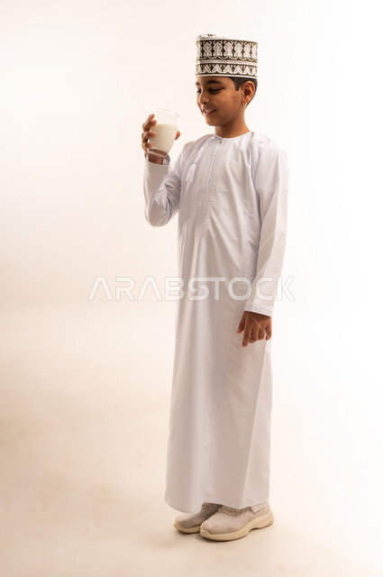 المحافظة على سلامة الجهاز المناعي، اتباع نظام غذائي صحي، فوائد مشتقات اللبن المتعددة للجسم والعظام, بورتريه لولد عربي خليجي عماني يرتدي الدشداشة والكمة يقوم بشرب كأس من الحليب، مفهوم العناية والاهتمام بالصحة, الجسم بالطول الكامل، خلفية بيضاء