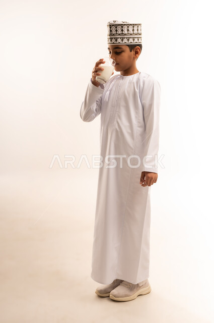 فوائد مشتقات اللبن المتعددة للجسم والعظام, بورتريه لولد عربي خليجي عماني يرتدي الدشداشة والكمة يقوم بشرب كأس من الحليب، المحافظة على سلامة الجهاز المناعي، مفهوم العناية والاهتمام بالصحة, اتباع نظام غذائي صحي، الجسم بالطول الكامل، خلفية بيضاء