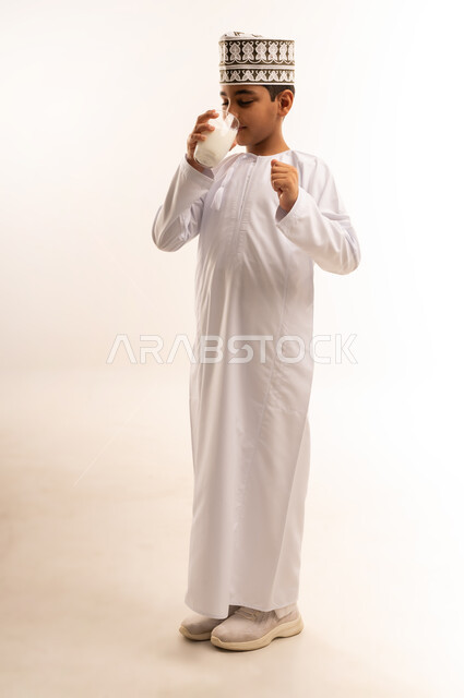 اتباع نظام غذائي صحي، فوائد مشتقات اللبن المتعددة للجسم والعظام, بورتريه لولد عربي خليجي عماني يرتدي الدشداشة والكمة يقوم بشرب كأس من الحليب، المحافظة على سلامة الجهاز المناعي، مفهوم العناية والاهتمام بالصحة, الجسم بالطول الكامل، خلفية بيضاء