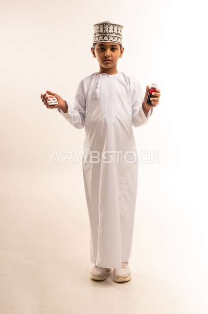 مفهوم الاستفادة من الدواء الموصوف, بورتريه لفتى عربي خليجي عماني يرتدي الدشداشة والكمة يحمل زجاجة دواء عاتمة وبرشام، الإصابة بالمرض والتوعك, الالتزام باتخاذ الأدوية في الوقت المناسب لها, صورة الجسم بالطول الكامل, خلفية بيضاء