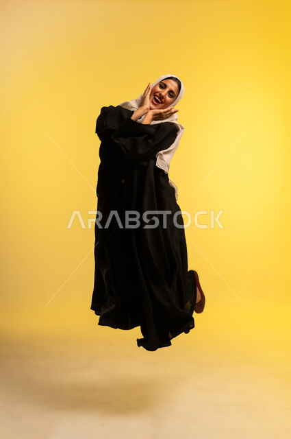 الاهتمام بالمظهر الخارجي، مفهوم الأنوثة والنعومة، شابة سعودية تقفز عاليا بايماءات الفرح والسرور، بورتريه لامرأة عربية خليجية عمانية ترتدي الحجاب والعباءة تضع يديها على خديها تنظر إلى الكاميرا بتعابير السعادة, الجسم بالطول الكامل، خلفية صفراء
