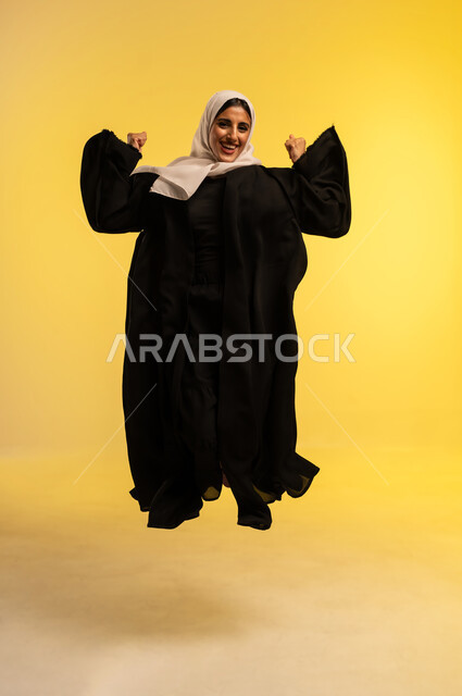 Concern about appearance, concept of femininity and softness, young Saudi woman jumping high with gestures of joy and happiness, portrait of an Arab Gulf Omani woman wearing hijab and abaya raising her hands looking at the camera with expressions of happiness, full body, yellow background