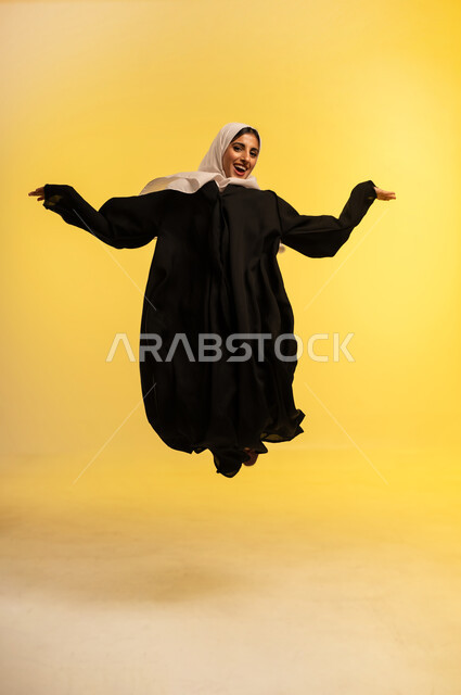 مفهوم الأنوثة والنعومة، شابة سعودية تقفز عاليا بايماءات الفرح والسرور، بورتريه لامرأة عربية خليجية عمانية ترتدي الحجاب والعباءة ترفع يديها تنظر إلى الكاميرا بتعابير السعادة, الاهتمام بالمظهر الخارجي، الجسم بالطول الكامل، خلفية صفراء