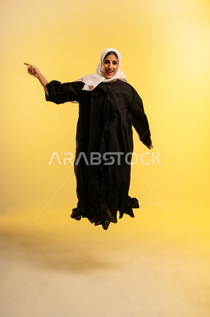 الاشارة باصبع السبابة إلى شيء ما، مفهوم الأنوثة والنعومة، الاهتمام بالمظهر الخارجي، شابة سعودية تقفز عاليا بايماءات الفرح والسرور، بورتريه لامرأة عربية خليجية عمانية ترتدي الحجاب والعباءة تنظر إلى الكاميرا بتعابير السعادة, الجسم بالطول الكامل، خلفية صفراء