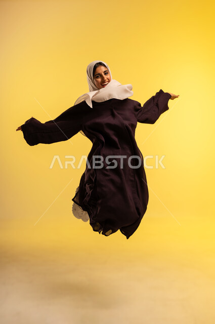 مفهوم الأنوثة والنعومة، شابة سعودية تقفز عاليا بايماءات الفرح والسرور، بورتريه لامرأة عربية خليجية عمانية ترتدي الحجاب والعباءة ترفع يديها تنظر إلى الكاميرا بتعابير السعادة, الاهتمام بالمظهر الخارجي، الجسم بالطول الكامل، خلفية صفراء
