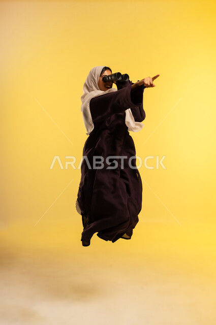 الاشارة باصبع السبابة إلى عنصر معين، شابة سعودية تقوم برؤية الأشياء البعيدة، التخطيط المستقبلي للأهداف والطموحات، بورتريه لامرأة عربية خليجية عمانية ترتدي العباءة والحجاب تقفز عاليا تمسك منظار ثنائي العينين تنظر إلى شيء ما، الجسم بالطول الكامل، خلفية صفراء