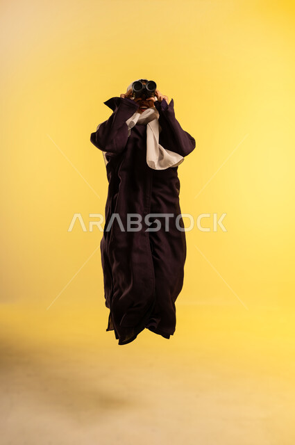 التفكير والتطلع الى المستقبل، شابة سعودية تقوم برؤية الأشياء البعيدة، التخطيط المستقبلي للأهداف والطموحات، بورتريه لامرأة عربية خليجية عمانية ترتدي العباءة والحجاب تقفز عاليا تمسك منظار ثنائي العينين تنظر إلى شيء ما، الجسم بالطول الكامل، خلفية صفراء
