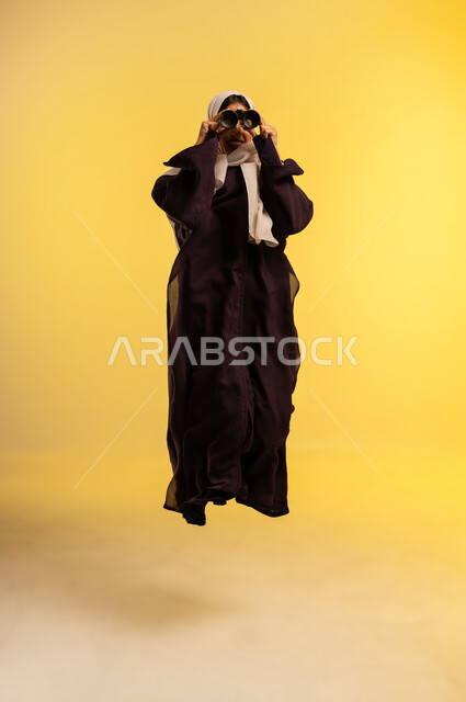 التخطيط المستقبلي للأهداف والطموحات، حب الاستكشاف والمغامرة، شابة سعودية تقوم برؤية الأشياء البعيدة، بورتريه لامرأة عربية خليجية عمانية ترتدي العباءة والحجاب تقفز عاليا تمسك منظار ثنائي العينين تنظر إلى شيء ما، الجسم بالطول الكامل، خلفية صفراء