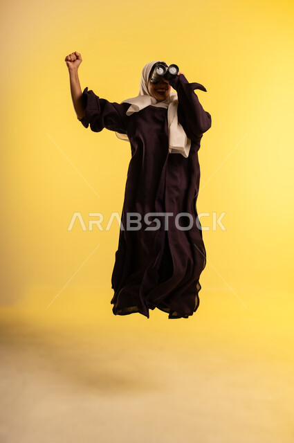 التخطيط المستقبلي للأهداف والطموحات، حب الاستكشاف والمغامرة، شابة سعودية تقوم برؤية الأشياء البعيدة، بورتريه لامرأة عربية خليجية عمانية ترتدي العباءة والحجاب تقفز عاليا تمسك منظار ثنائي العينين تنظر إلى شيء ما، الجسم بالطول الكامل، خلفية صفراء