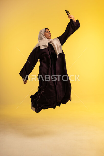 الدردشة مع الاصدقاء عبر الجوال، استخدام جهاز تقني حديث، بورتريه لامرأة عربية خليجية عمانية ترتدي العباءة والحجاب تقوم بإجراء مكالمة فيديو عبر الهاتف المحمول تقفز عاليا بإيماءات الفرح لسماع اخبار جيدة، شابة سعودية تلتقط صورة سيلفي، الجسم بالطول الكامل، خلفية صفراء