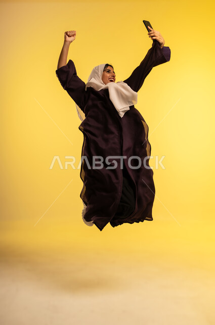 استخدام جهاز تقني حديث، بورتريه لامرأة عربية خليجية عمانية ترتدي العباءة والحجاب تقوم بإجراء مكالمة فيديو عبر الهاتف المحمول تقفز عاليا بإيماءات الفرح لسماع اخبار جيدة، الدردشة مع الاصدقاء عبر الجوال، شابة سعودية تلتقط صورة سيلفي، الجسم بالطول الكامل، خلفية صفراء